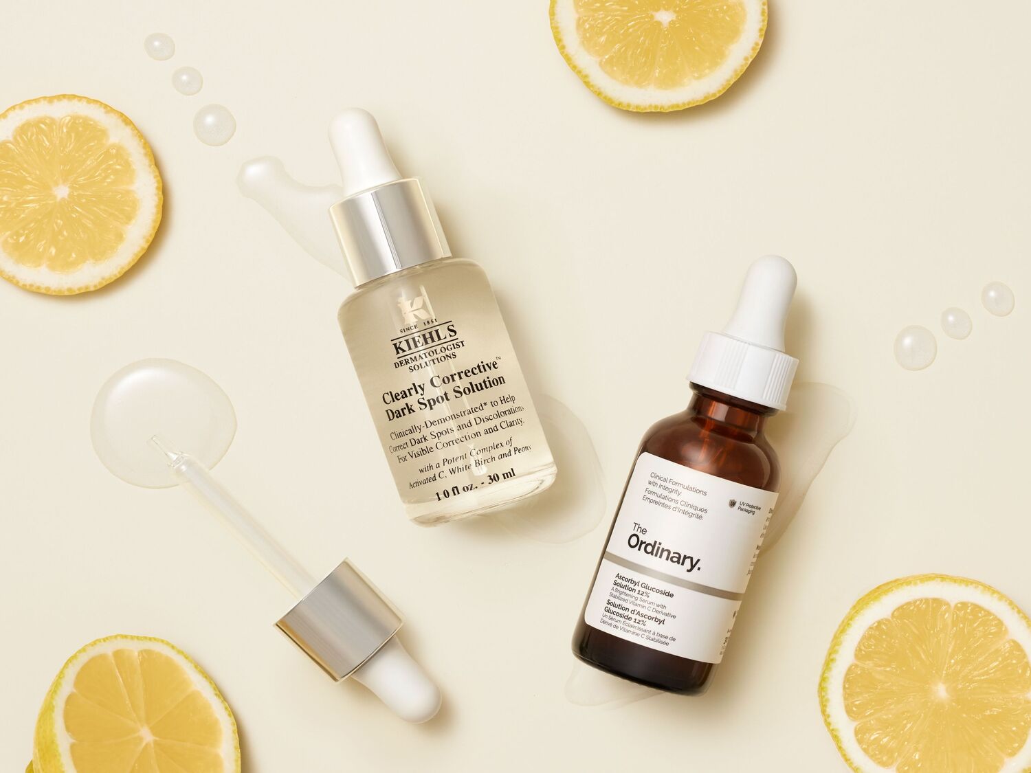 Deux sérums éclaircissants, Kiehl's Clearly Corrective Dark Spot Solution et The Ordinary Ascorbyl Glucoside Solution 12%, sont présentés avec des tranches de citron et des gouttes de produit sur un fond clair.