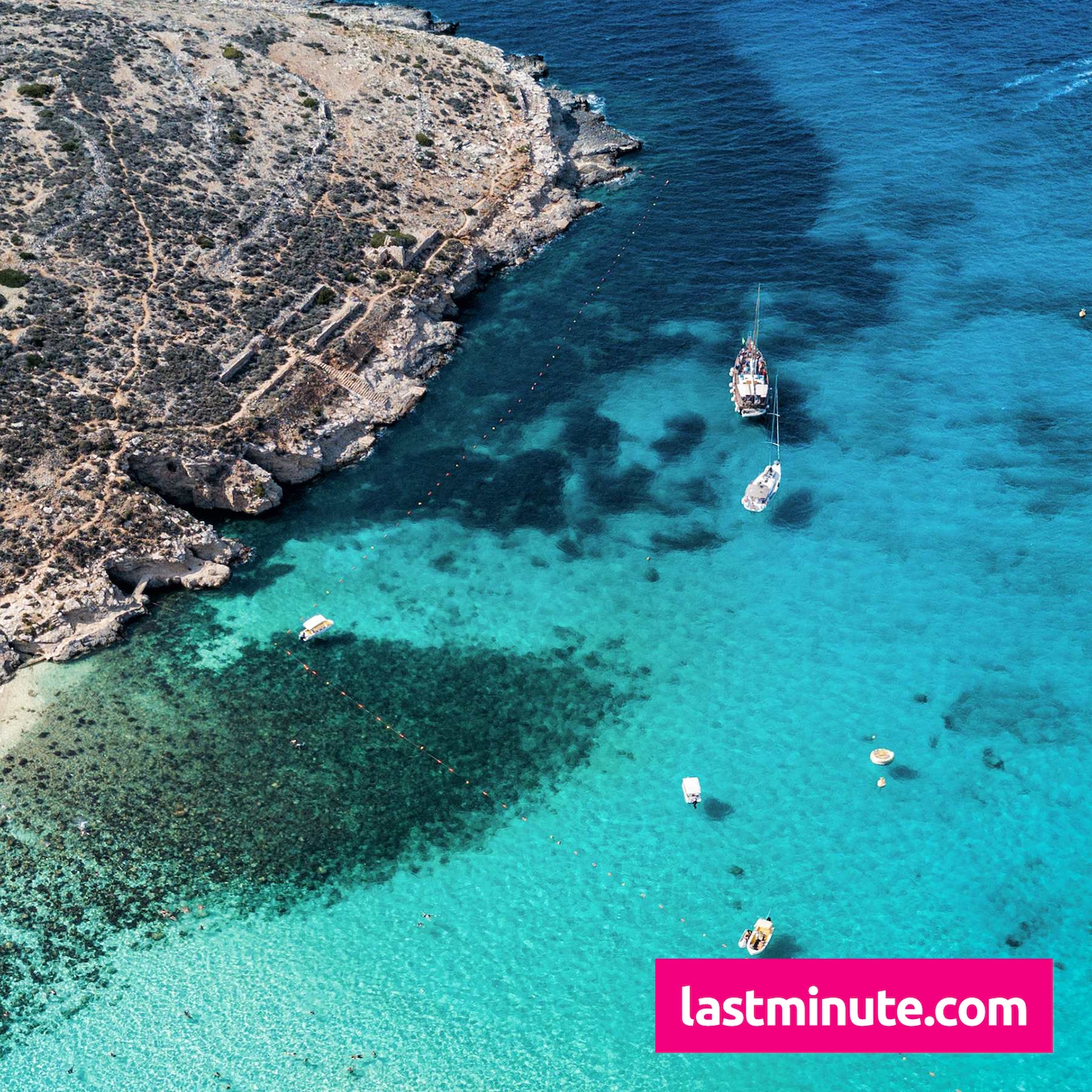 Ein Luftbild zeigt eine felsige Küste mit kargem Land und türkisfarbenem Wasser, in dem mehrere Boote und Schwimmer zu sehen sind, mit einem rosa Banner von lastminute.com am unteren Rand.