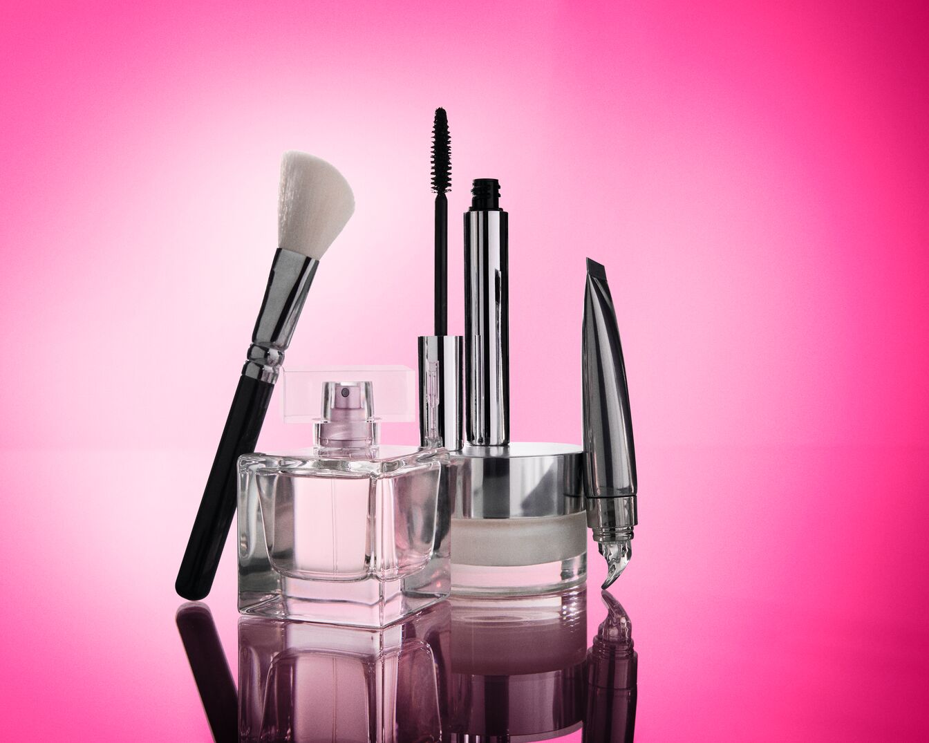 Eine Sammlung von Make-up-Produkten, darunter ein Pinsel, Parfüm, Mascara, ein Tiegel mit Creme und ein Augenbrauenstift, ist auf einem leuchtend pinken Hintergrund angeordnet.