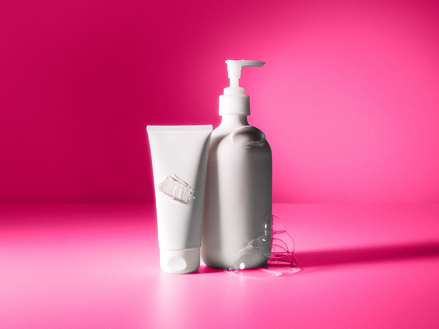 Ein weißer Tubenbehälter mit einem cremefarbenen Klecks und eine weiße Pumpspenderflasche mit klaren Blasen stehen auf einem leuchtend pinken Hintergrund.