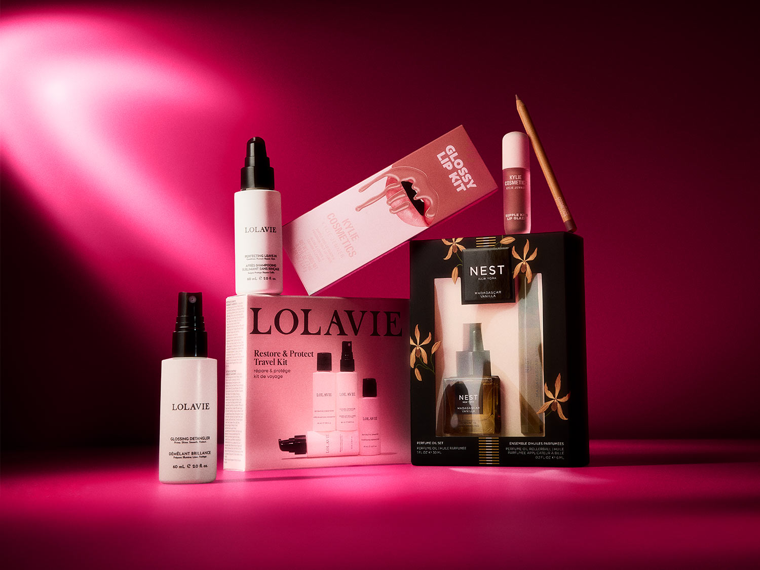 Das Bild zeigt eine Auswahl an Schönheitsprodukten, darunter ein Lolavie Restore & Protect Travel Kit, ein Kylie Cosmetics Glossy Lip Kit und ein Nest New York Madagascar Vanilla Perfume Oil Set, die alle auf einem leuchtend magentafarbenen Hintergrund angeordnet sind.