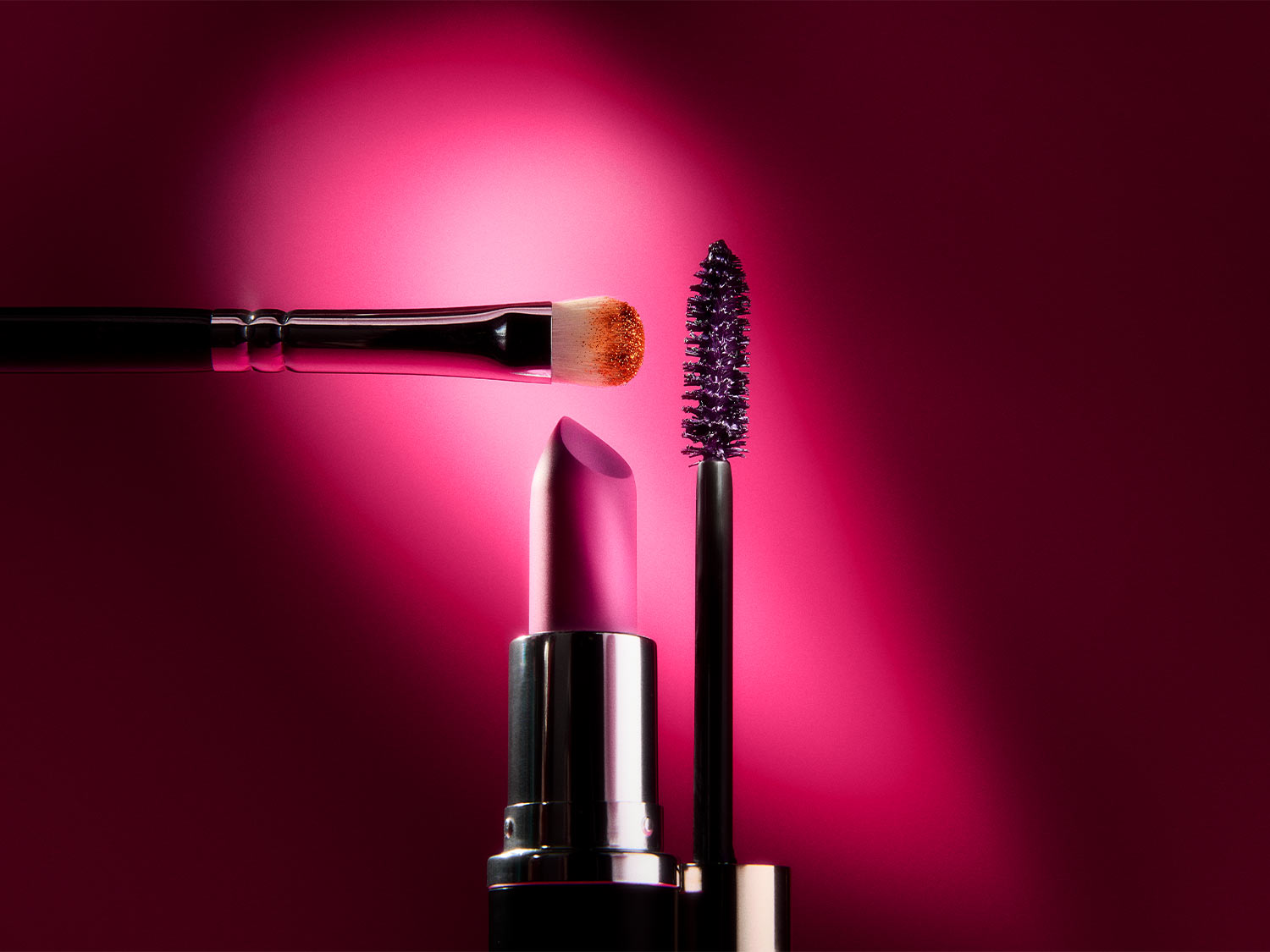 Ein rosafarbener Lippenstift, eine Mascarabürste mit lila Mascara und ein Make-up-Pinsel mit goldenem Lidschatten sind vor einem leuchtend rosafarbenen Hintergrund angeordnet.