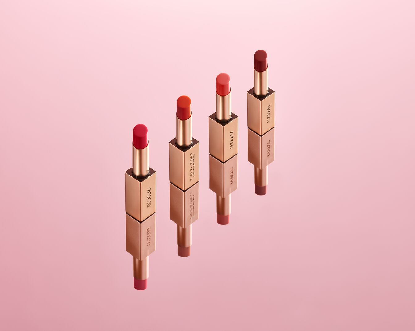 Vier Douglas 2-in-1 lippenbalsems in verschillende tinten rood en oranje, elk met een goudkleurige verpakking, staan rechtop op een roze achtergrond.