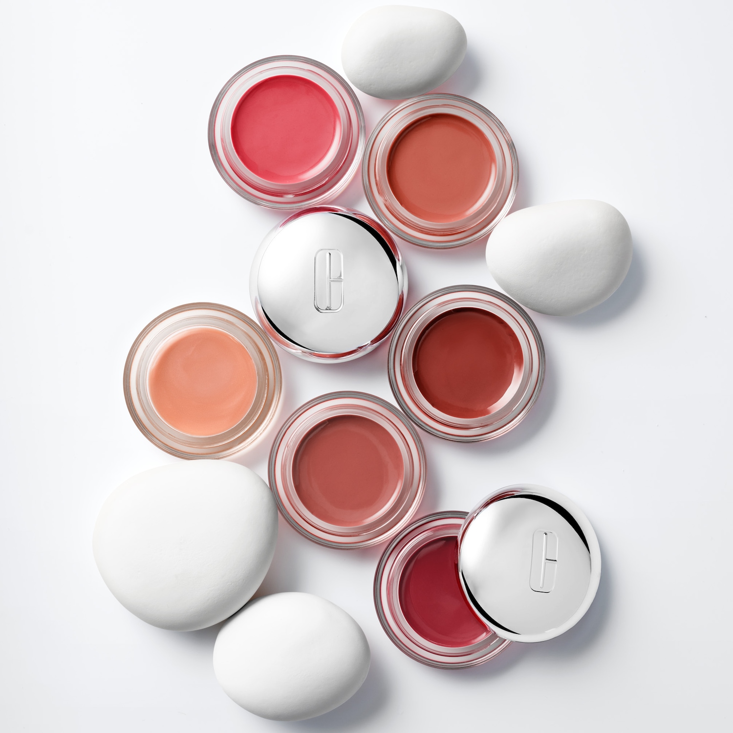 Une sélection de baumes Clinique Daily Calm Lip + Cheek Color Balms déclinés en différentes teintes, allant du rose tendre au rouge vif, présentés dans de petits pots ronds aux couvercles argentés, entourés de pierres blanches lisses.