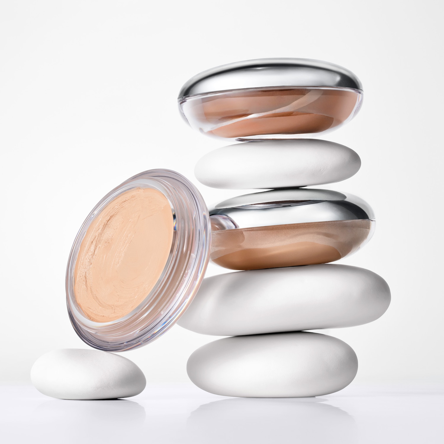 Deux boîtes à maquillage rondes et ouvertes, dotées de couvercles argentés et contenant un produit beige, sont posées sur une pile de pierres blanches et lisses ; l'une d'elles est légèrement inclinée vers la gauche.