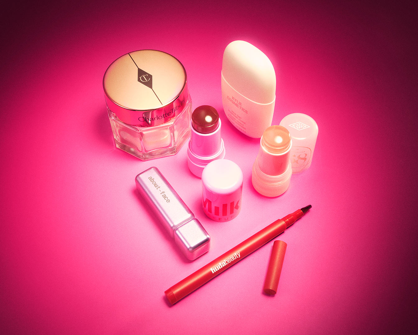 Eine Sammlung von Make-up-Produkten, darunter ein Charlotte Tilbury Tiegel, Kylie Cosmetics und Milk Makeup Produkte, ein About-Face Lippenstift und ein Huda Beauty Eyeliner, die auf einem leuchtend pinkfarbenen Hintergrund angeordnet sind.
