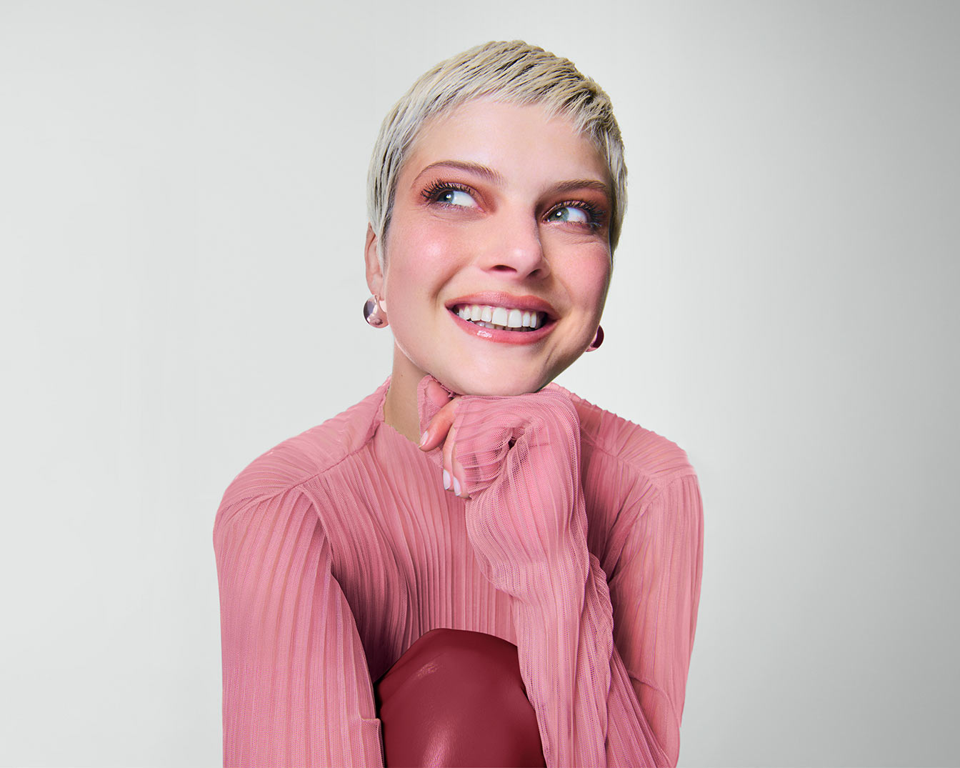 Eine junge Frau mit kurzen blonden Haaren, die ein rosafarbenes, plissiertes Oberteil trägt und lächelnd zur Seite blickt, während sie ihr Kinn auf ihre Hand stützt.