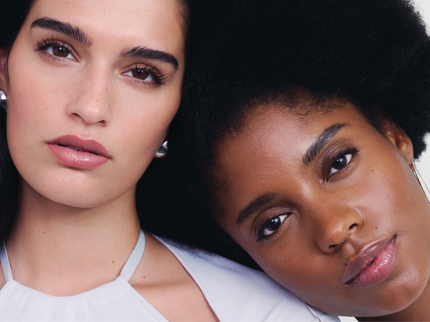 Due donne, una con capelli scuri e l'altra con capelli afro, mostrano labbra lucide e carnose, con la donna dai capelli scuri che indossa orecchini a bottone argentati.