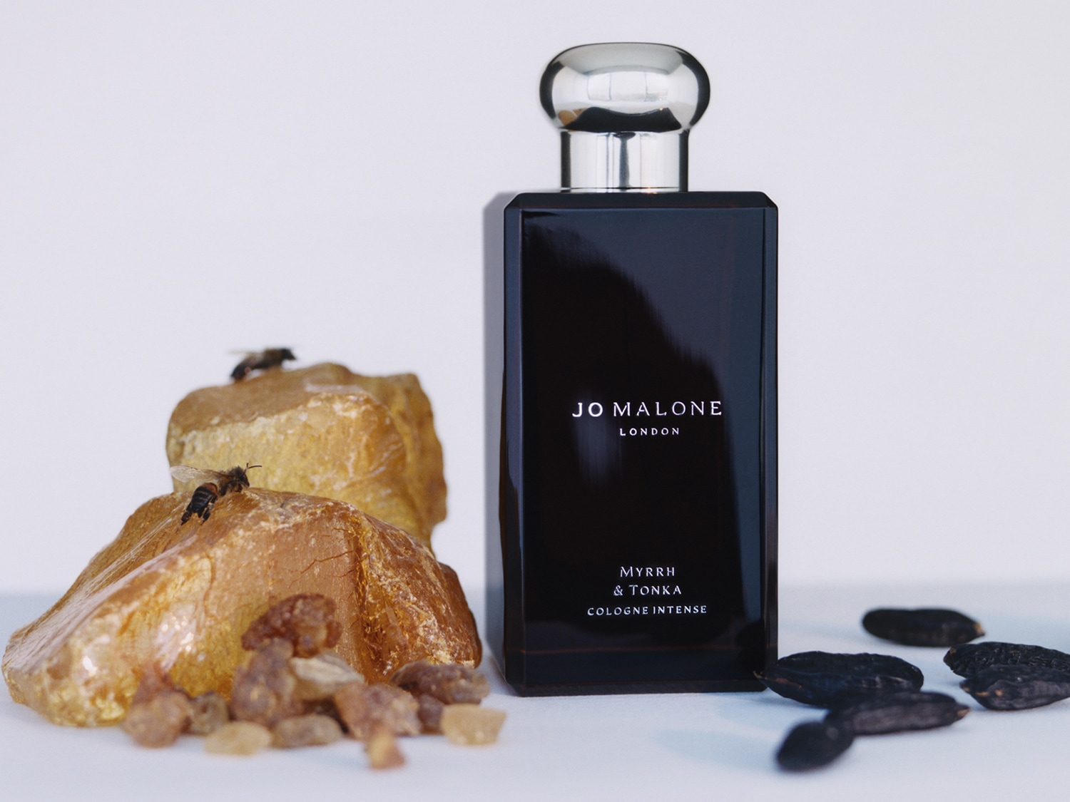 Il profumo Jo Malone London Myrrh & Tonka Cologne Intense in una bottiglia nera lucida con tappo argentato, affiancato da ambra grezza, grani di mirra e fave di tonka, con due api che si posano sull'ambra.