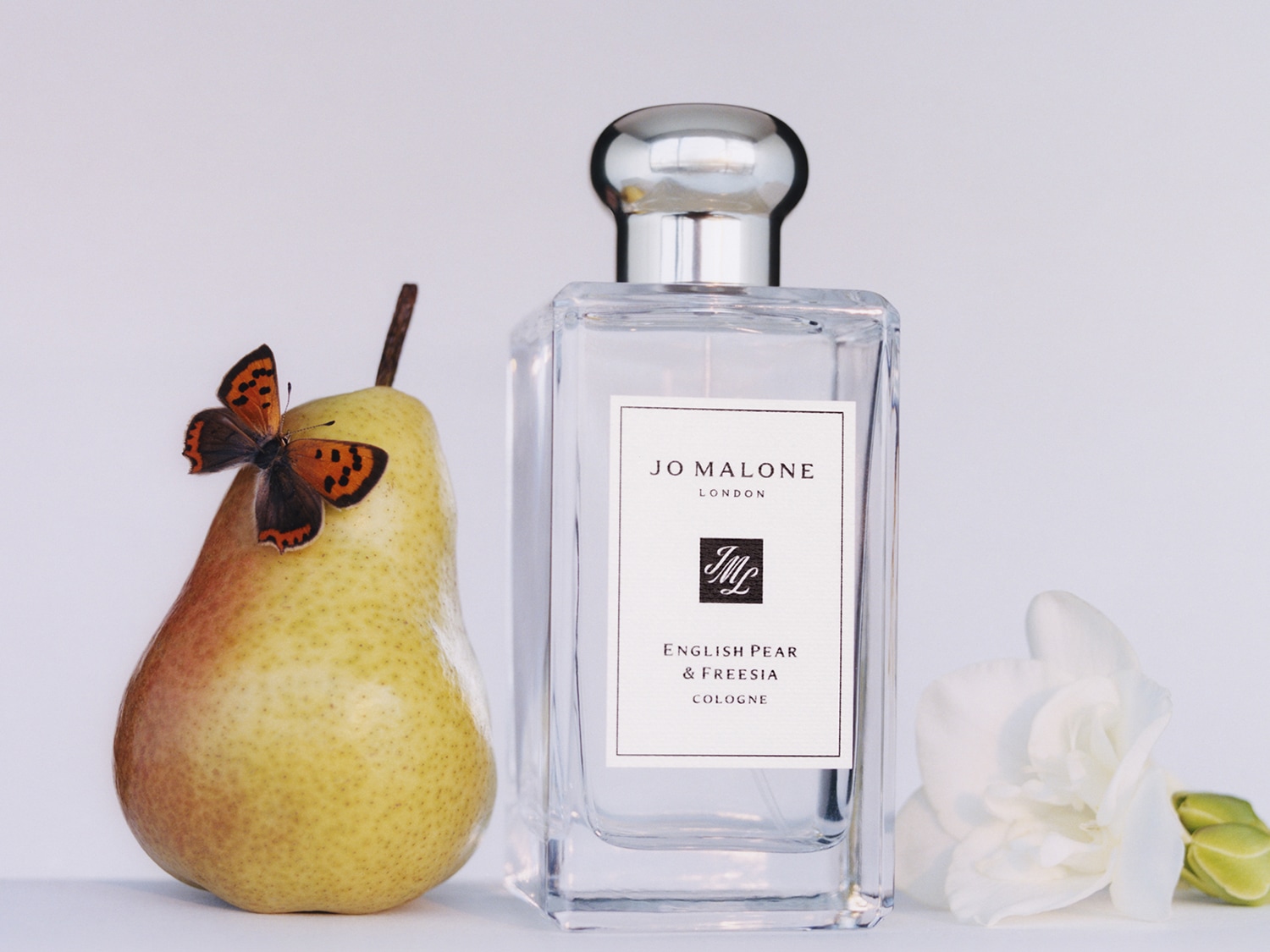 Un flacone trasparente di Jo Malone London English Pear & Freesia Cologne con tappo argentato, affiancato da una pera gialla con una farfalla arancione e un fiore bianco, su sfondo bianco.
