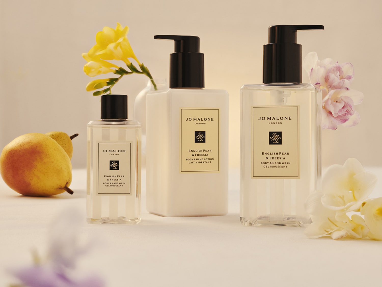 Tre prodotti Jo Malone London English Pear & Freesia, tra cui una lozione corpo e due bagnoschiuma, sono esposti con pere fresche e fiori bianchi e gialli, creando un'atmosfera elegante e naturale.