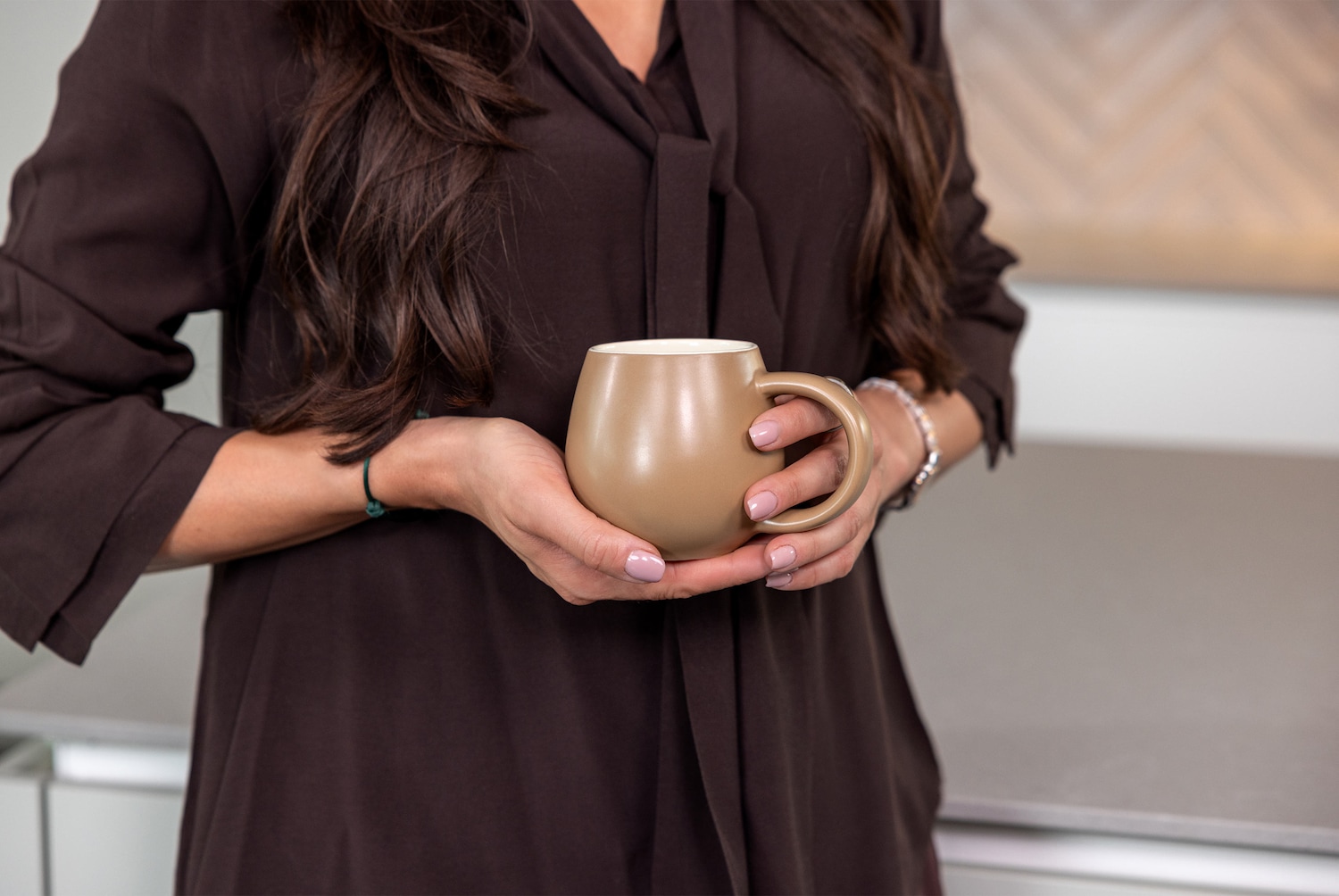 Eine Frau mit langen, braunen Haaren und einem dunkelbraunen Oberteil hält eine beigefarbene Tasse mit beiden Händen, ihre Nägel sind hellrosa lackiert.