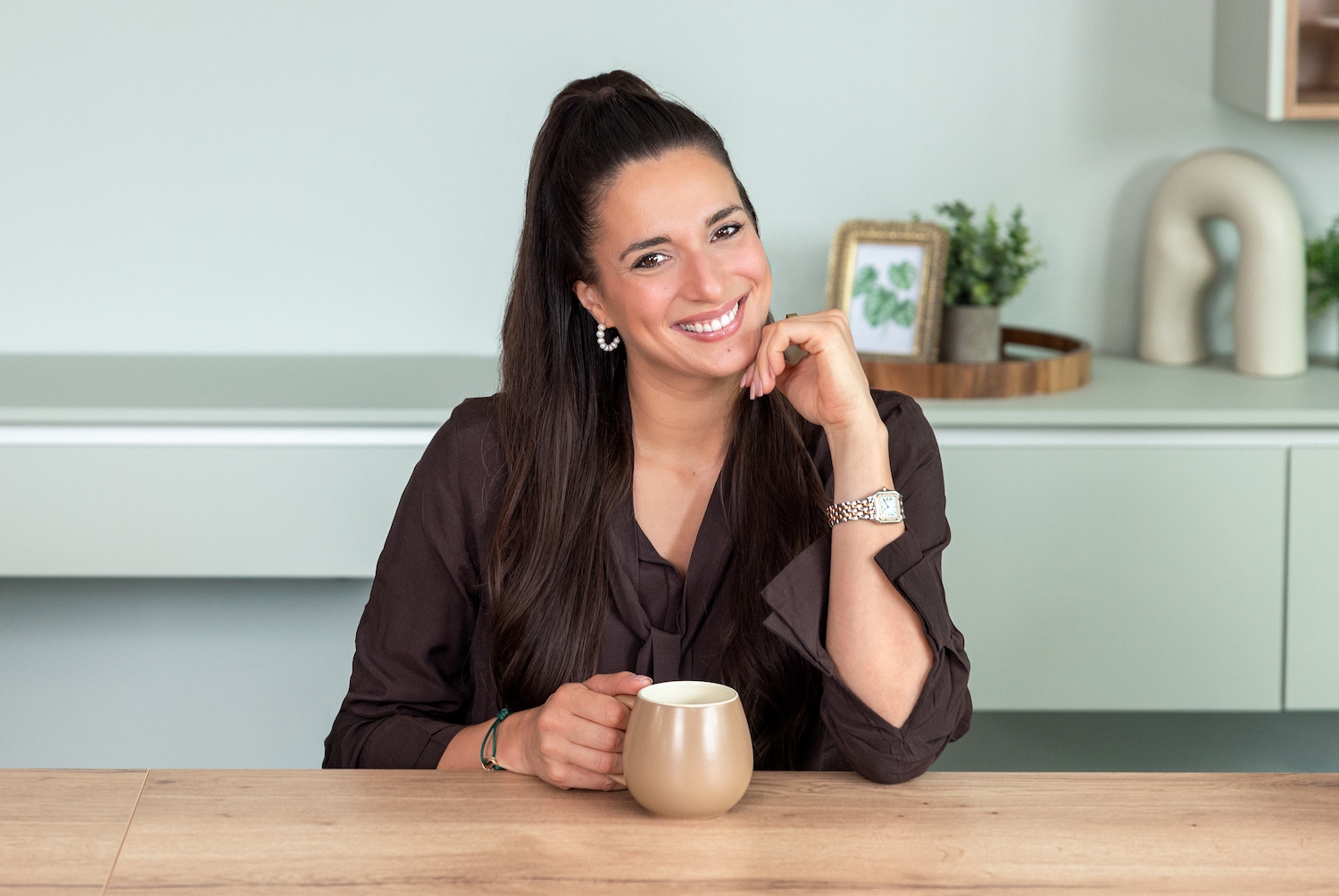 Eine lächelnde Frau mit langen, dunklen Haaren, die zu einem hohen Pferdeschwanz gebunden sind, sitzt an einem Holztisch und hält eine beigefarbene Tasse, während sie ihren Kopf auf ihre Hand stützt.