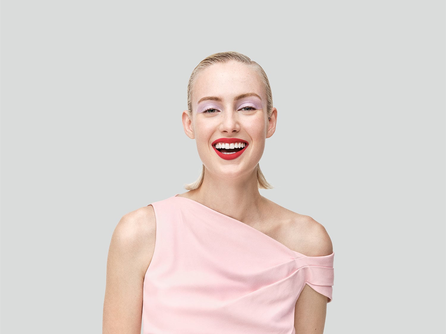 Eine strahlende Frau mit glattem blondem Haar trägt ein zartrosa Oberteil, leuchtend roten Lippenstift und sanften lila Lidschatten.