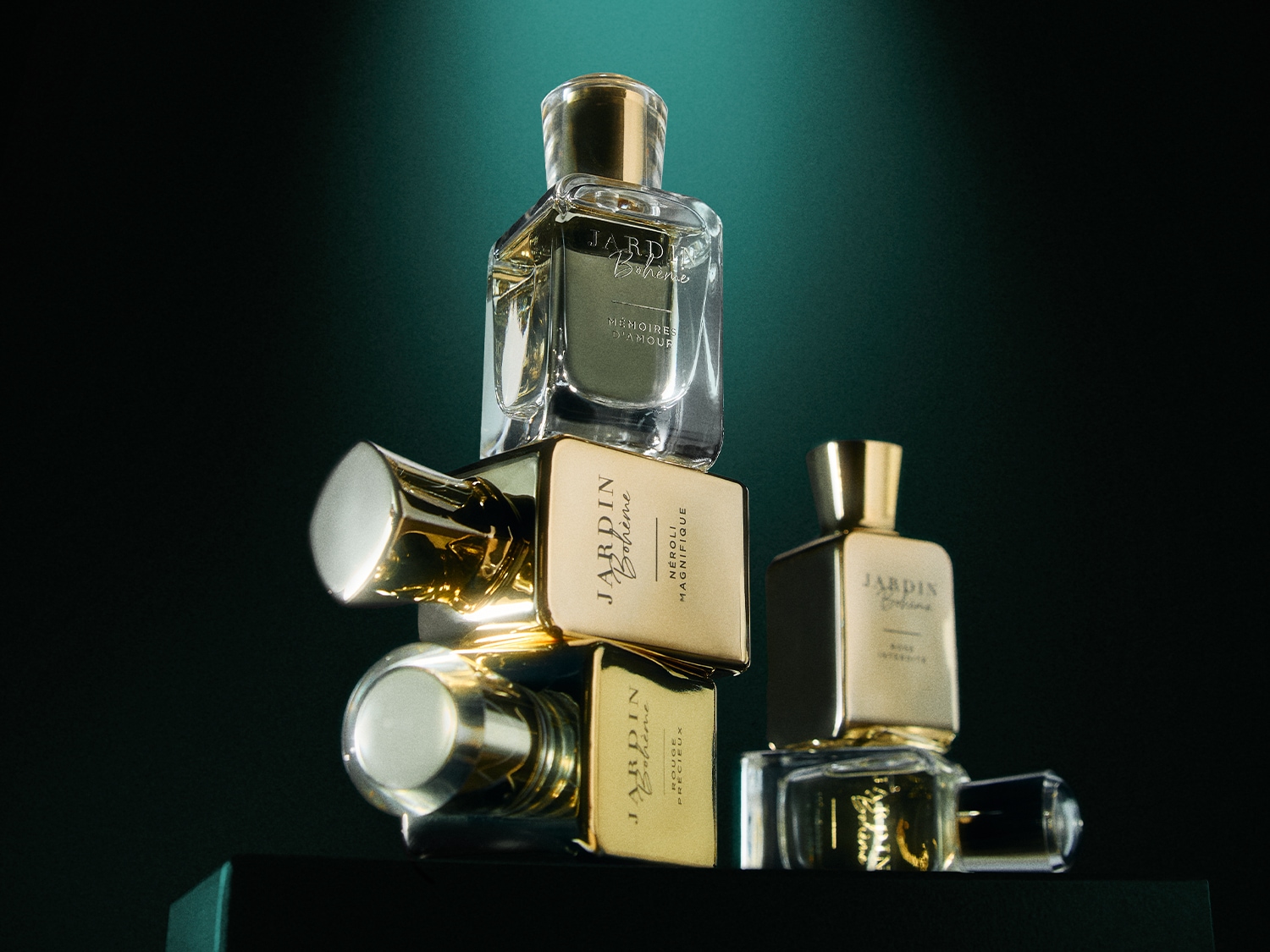 Drie parfumflesjes van Jardin Bohème, twee met gouden doppen en één met een transparante dop, staan opgestapeld tegen een donkergroene achtergrond.