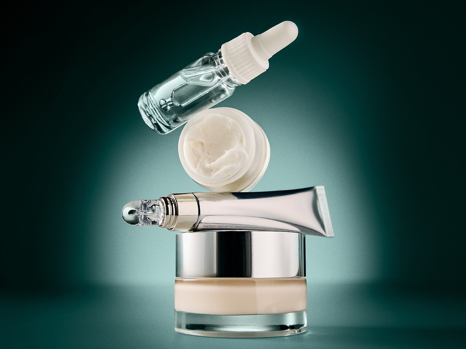 Een verzameling huidverzorgingsproducten, waaronder een serumflesje, een crème in een potje, een tube met een rollerapplicator en een foundationpotje, allemaal gestapeld tegen een donkergroene achtergrond.