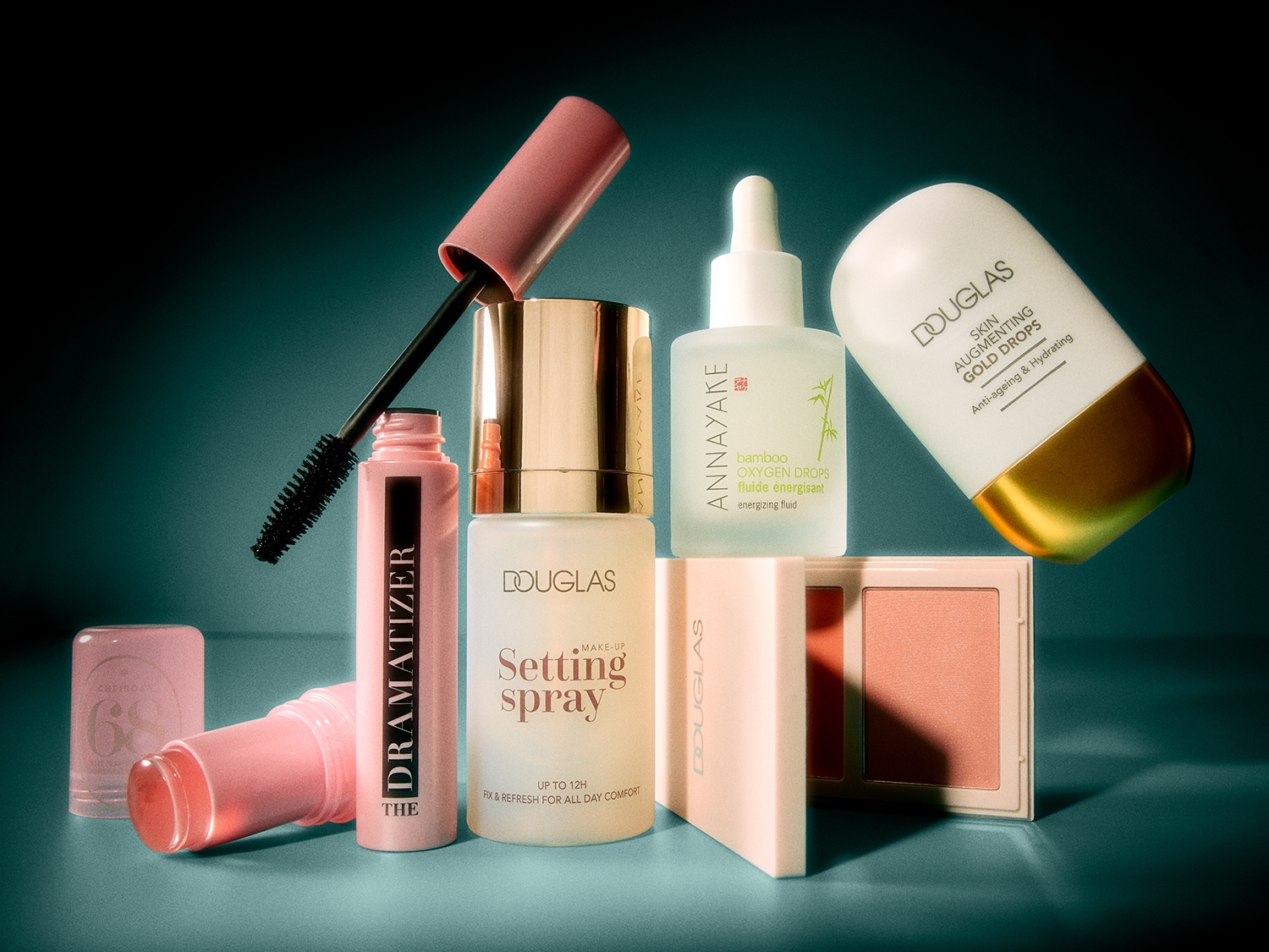Een roze mascara met een open dop en een zwart borsteltje, een roze lippenbalsem, een transparante setting spray, een Annayake Bamboo Oxygen Drops serum en een Douglas Skin Augmenting Gold Drops serum, samen met een Douglas blush in een lichtroze tint.