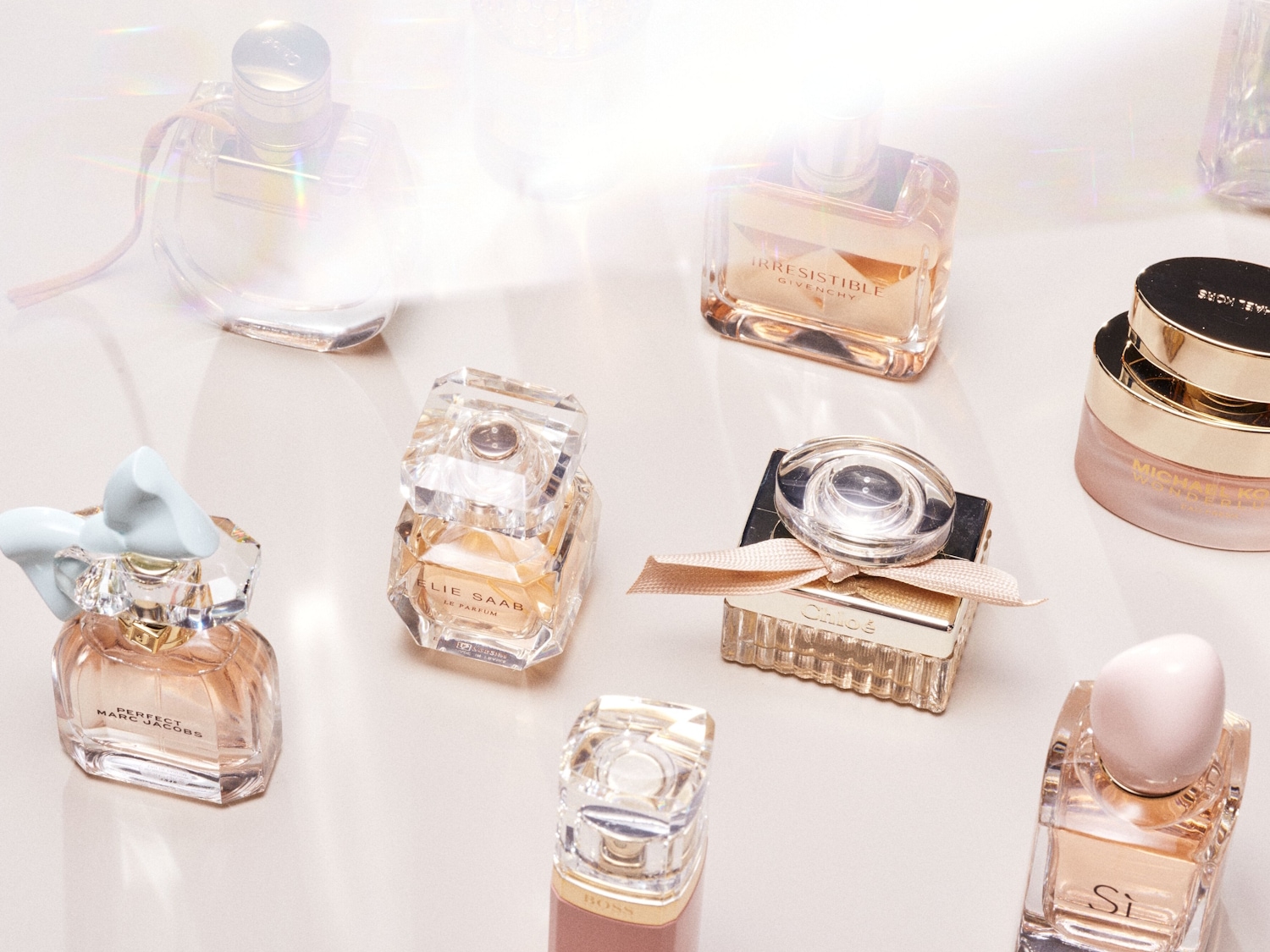 Eine Sammlung von Parfümflaschen, darunter Marc Jacobs Perfect mit einer hellblauen Schleife, Elie Saab Le Parfum in einem facettierten Flakon und Givenchy Irresistible in einem rechteckigen Flakon, alle auf einer hellen Oberfläche.