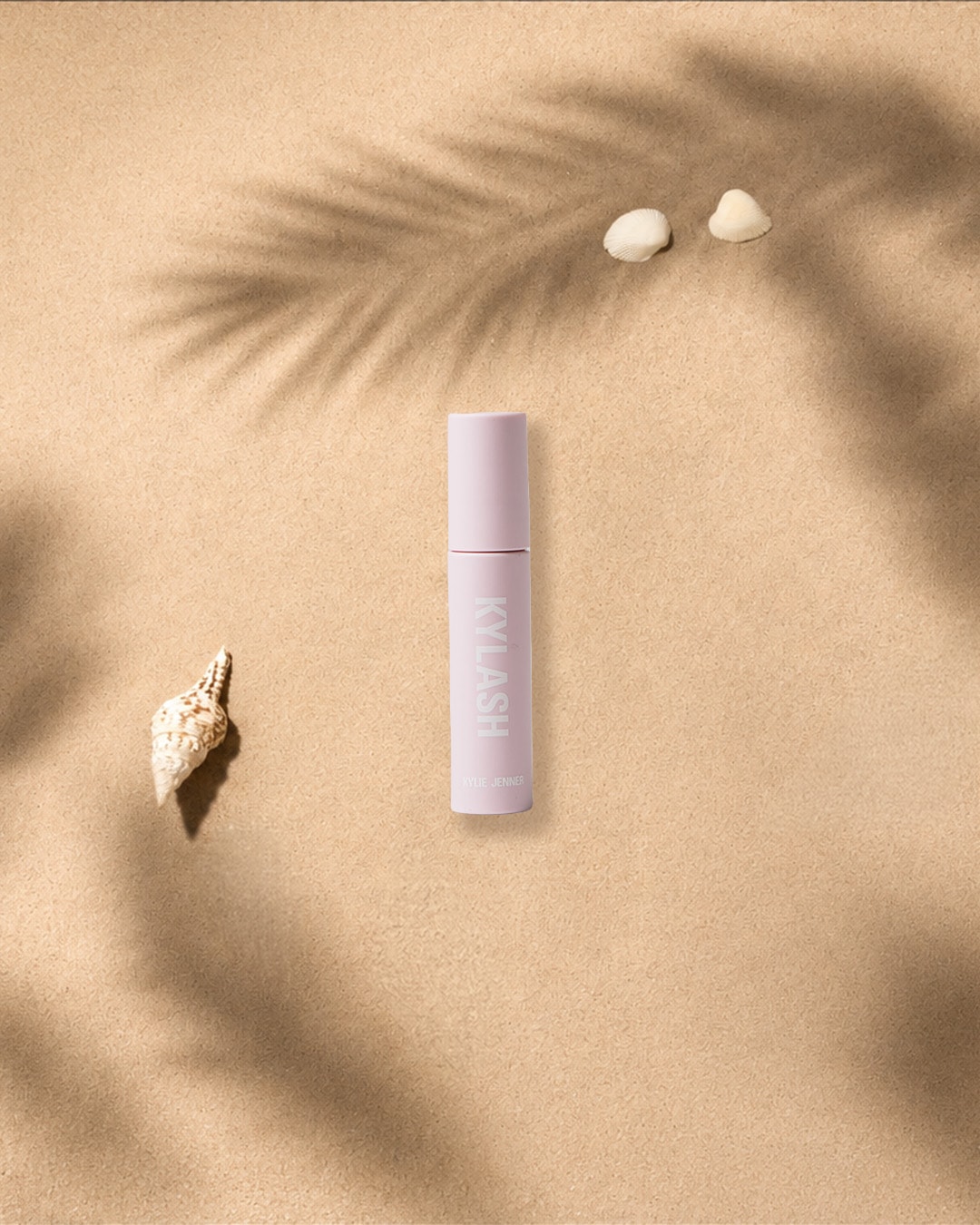 Die Kylie Jenner Kylash Mascara in einer hellvioletten Tube liegt auf beigem Sand, umgeben von Muscheln und Palmenblattschatten, die eine sommerliche Atmosphäre schaffen.