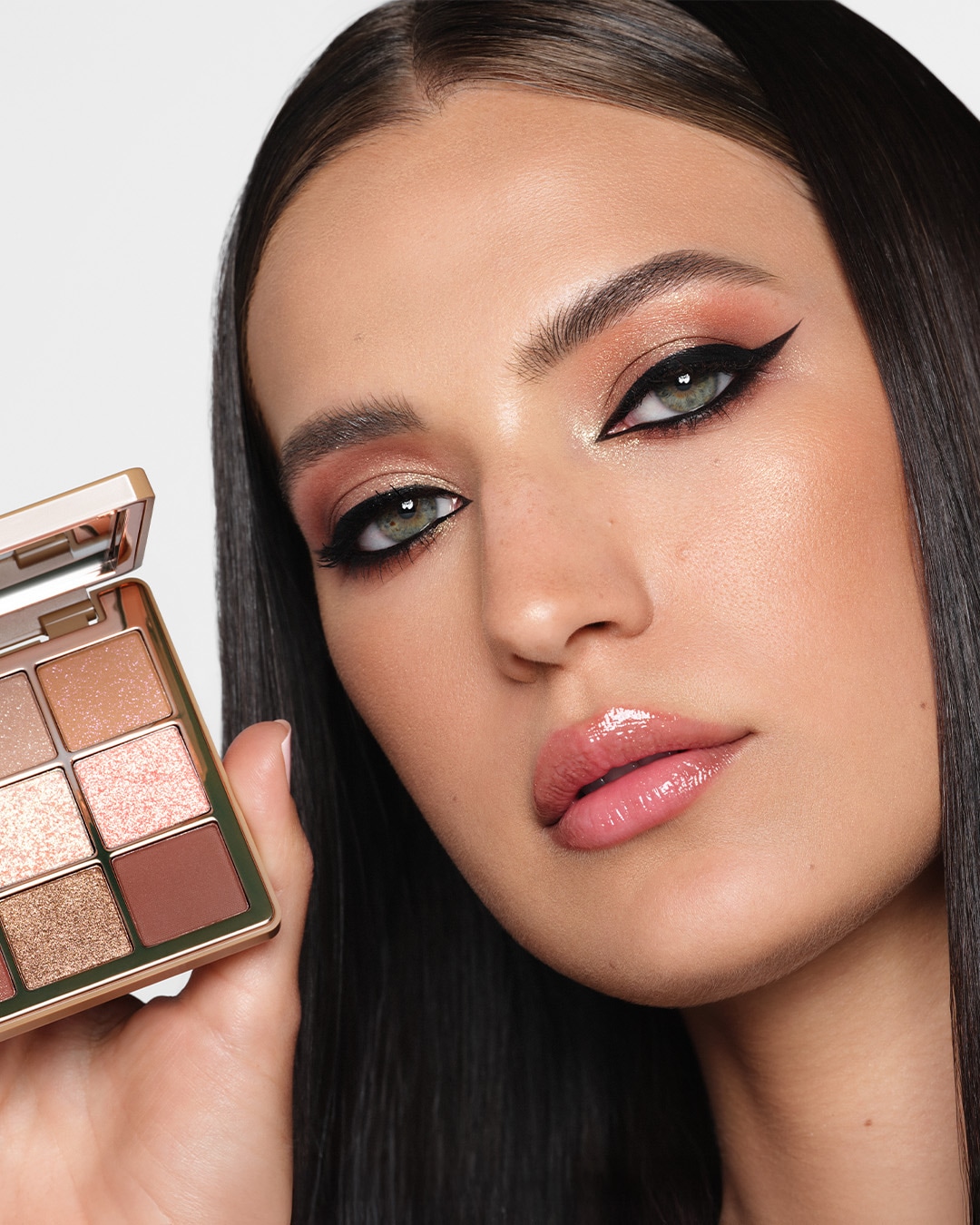 Un model cu părul negru, lung și drept, poartă un machiaj dramatic, cu eyeliner negru, lucios, și fard de pleoape roz-auriu, ținând în mână o paletă de farduri de pleoape cu șase nuanțe, în tonuri de maro și roz.