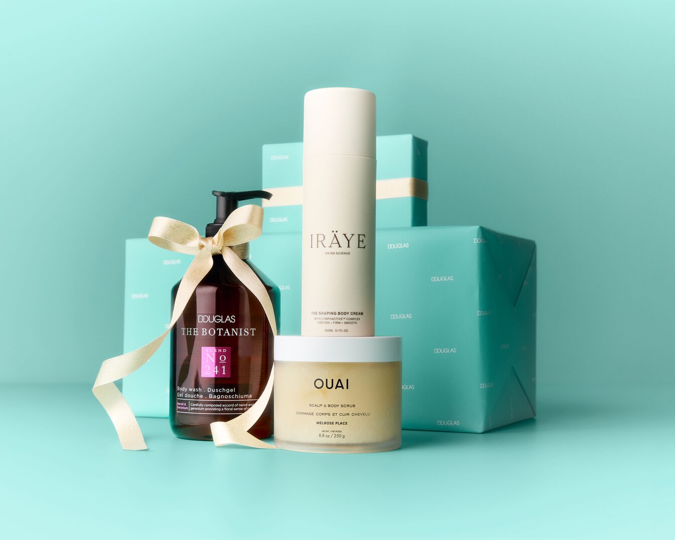 Un ensemble de produits de beauté Douglas, comprenant un gel douche The Botanist, une crème pour le corps Iräye et un gommage pour le cuir chevelu et le corps Ouai, est présenté sur un fond bleu sarcelle avec des boîtes cadeaux Douglas.