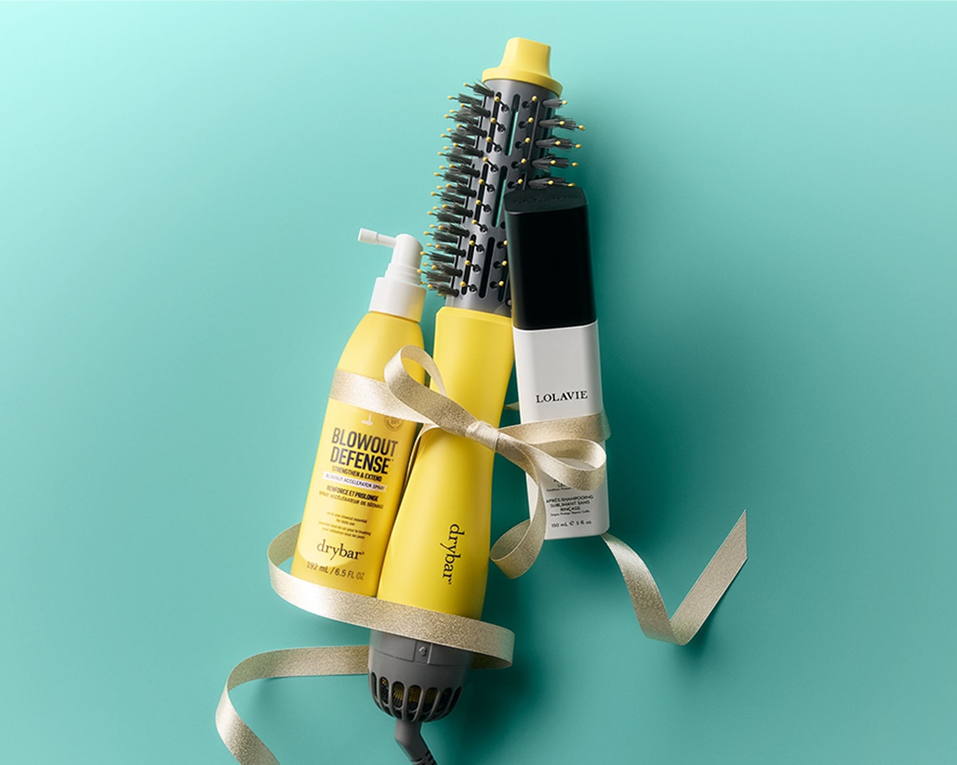 Un ensemble de produits capillaires Drybar et Lolavie, comprenant un spray Blowout Defense jaune et un spray après-shampooing blanc, est présenté avec une brosse à air chaud jaune et noire, le tout attaché par un ruban doré sur un fond bleu sarcelle.