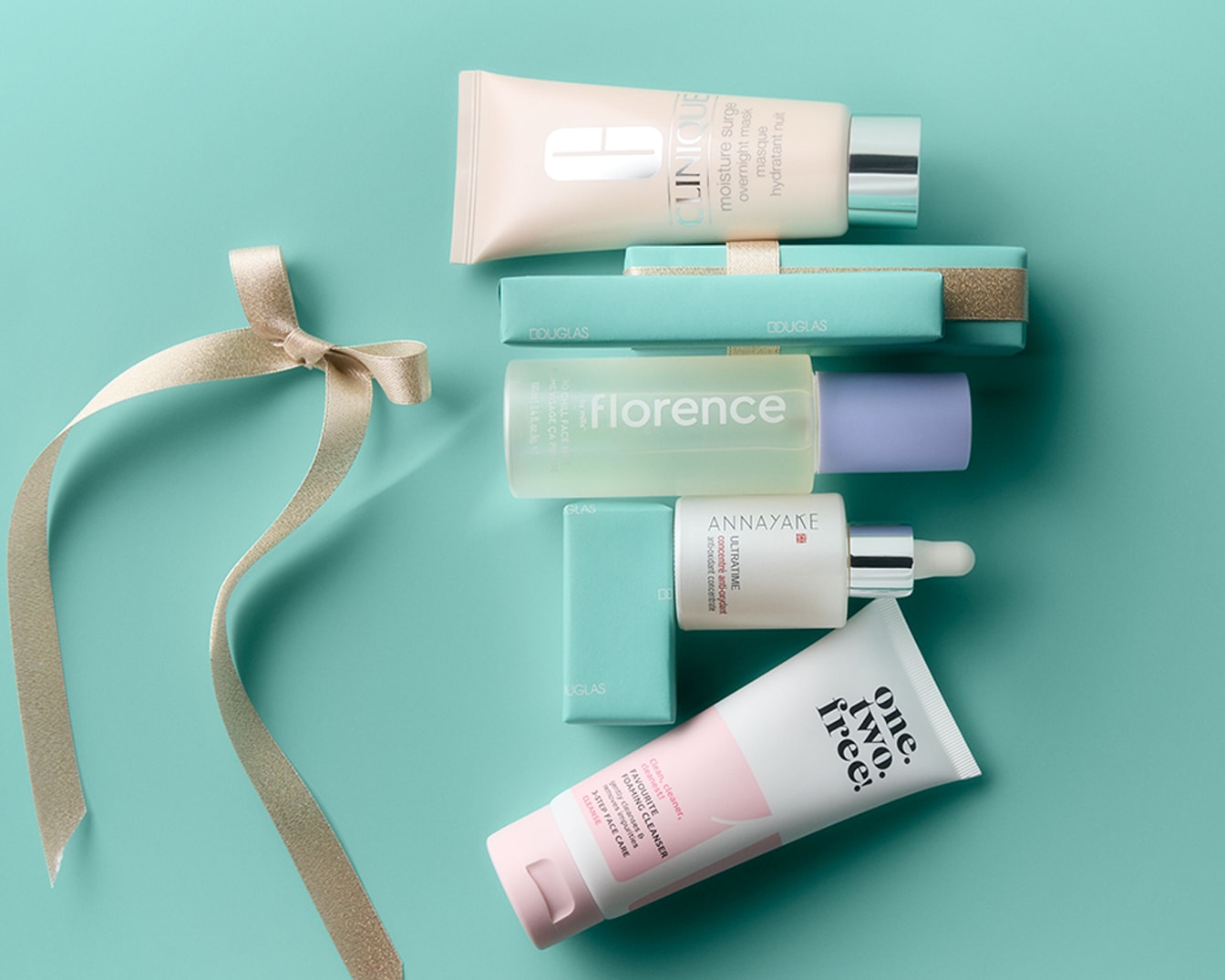 Découvrez une sélection de produits de soin pour le visage, incluant un masque de nuit Clinique, un sérum Annayake, un nettoyant moussant One.Two.Free! et un soin Florence, présentés avec un ruban doré sur un fond bleu-vert.