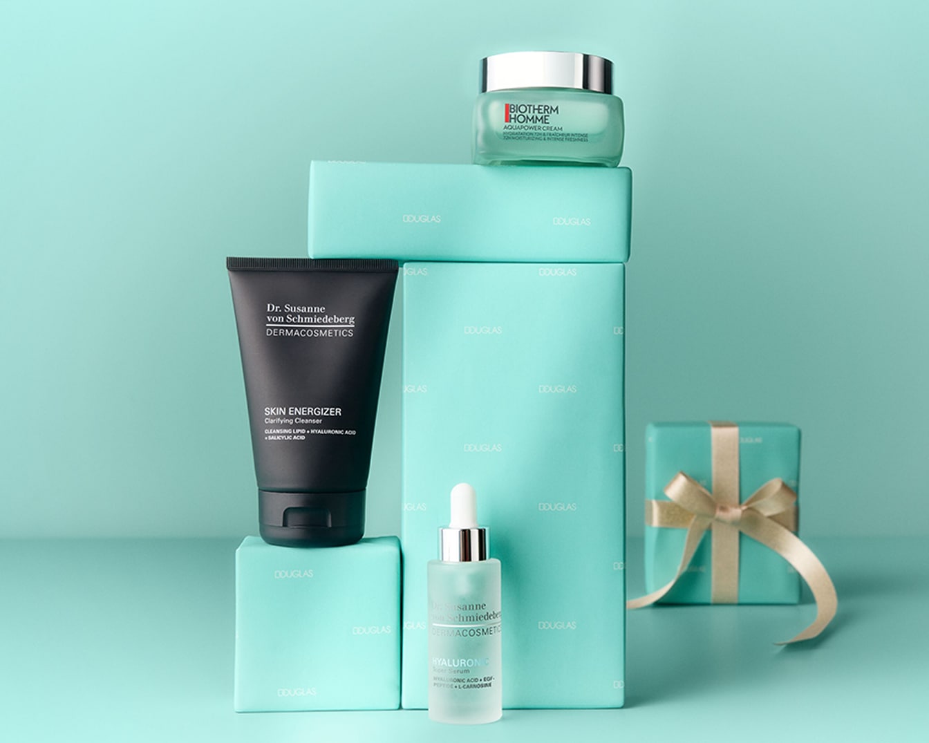 Ein schwarzer Dr. Susanne von Schmiedeberg Skin Energizer klärender Reiniger und ein Biotherm Homme Aquapower Creme stehen auf Douglas-Geschenkboxen, die auf einem hellblauen Hintergrund gestapelt sind.