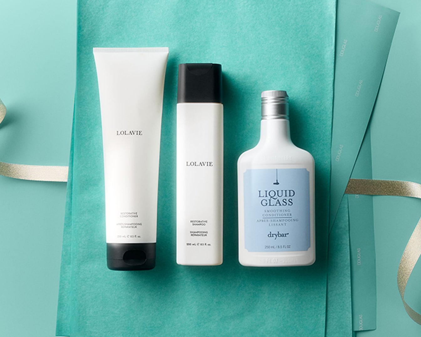 Drei Haarpflegeprodukte, darunter Lolavie Restorative Conditioner und Shampoo sowie Drybar Liquid Glass Smoothing Conditioner, sind auf einem türkisfarbenen Tuch mit einem goldenen Band arrangiert.