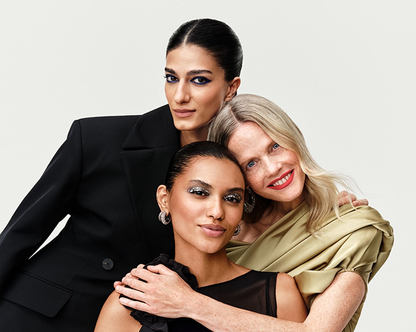 Drei Frauen mit unterschiedlichen Hauttönen und Haarfarben, die festlich geschminkt sind, posieren eng beieinander und lächeln in die Kamera.