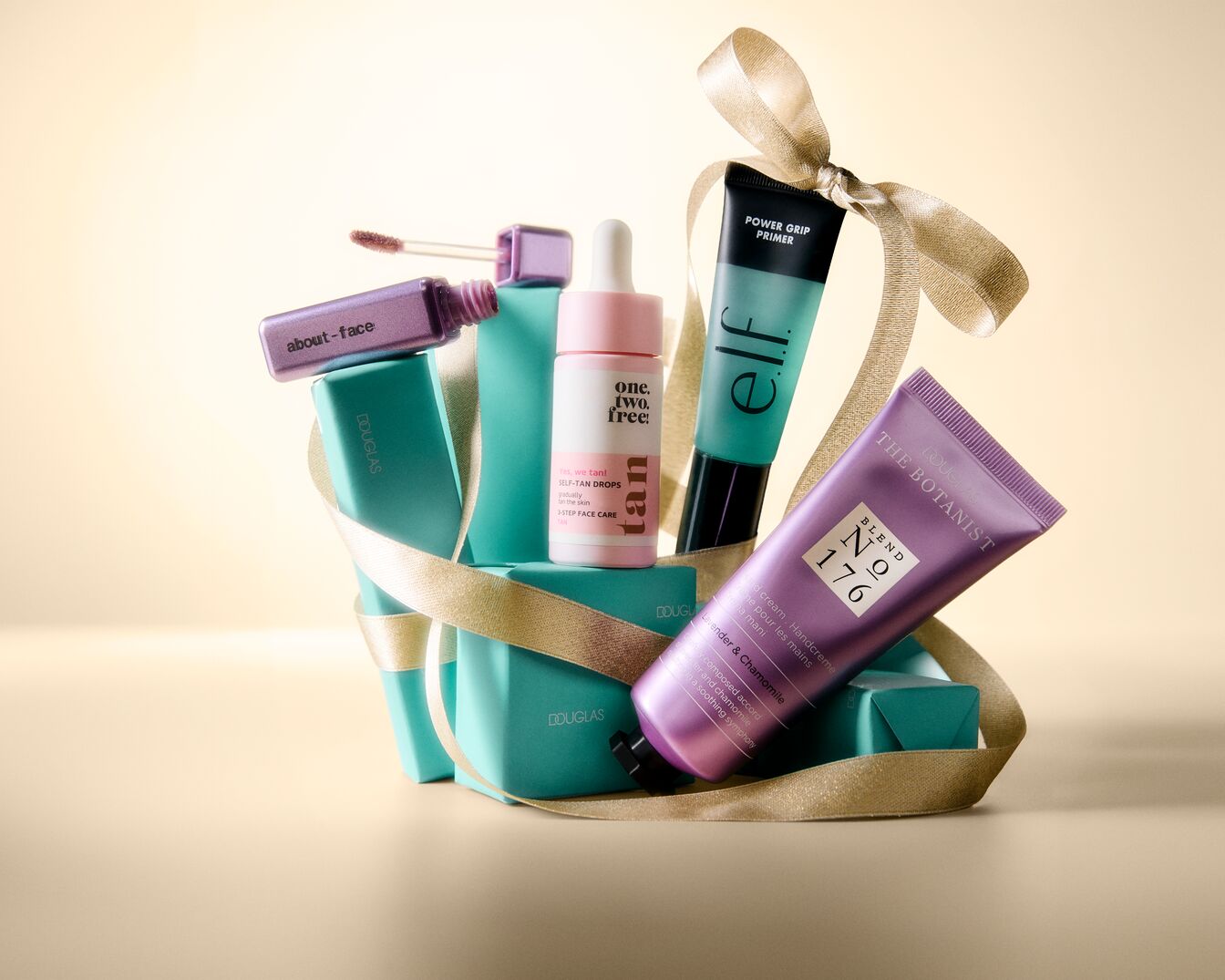 Ein festliches Arrangement aus Beauty-Produkten, darunter ein lila Lipgloss von "about-face", ein rosa Fläschchen "one.two.free!" Selbstbräunungstropfen, ein türkisfarbener "elf" Power Grip Primer und eine lila Handcreme "THE BOTANIST" von Douglas, alle umwickelt mit einem goldenen Band.