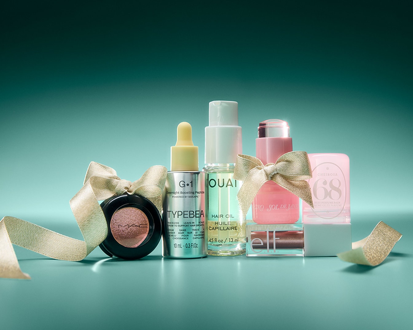 Eine Reihe von Schönheitsprodukten, darunter ein MAC Lidschatten, Typebea Overnight Boosting Peptide, Ouai Haaröl und Sol de Janeiro Cheirosa 68 Parfüm Balsam, sind festlich mit goldenen Bändern auf einem aquamarinfarbenen Hintergrund arrangiert.