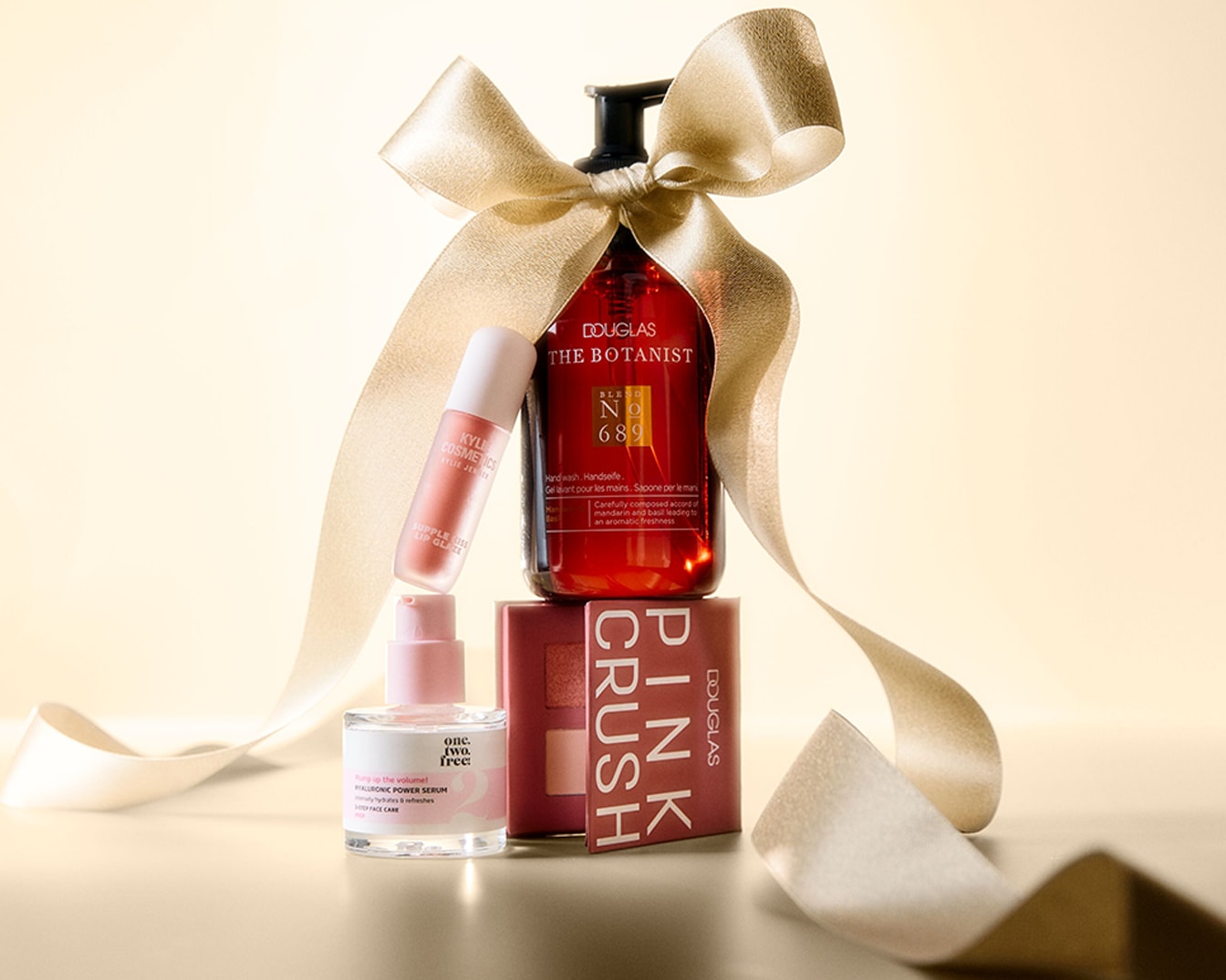 Een feestelijke geschenkset met een Douglas The Botanist handzeep, Kylie Cosmetics lipgloss, One.Two.Free. Hyaluronic Power Serum en een Douglas Pink Crush oogschaduwpalet, alles omwikkeld met een gouden lint.