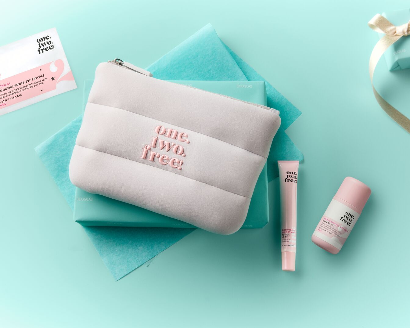 Een lichtroze make-up tasje met het logo "one. two. free!" in donkerroze letters, ligt op een turquoise ondergrond met twee turquoise geschenkdozen en een wit zakje met de tekst "HYALURONIC POWER EYE PATCHES" en "3-STEP FACE CARE".