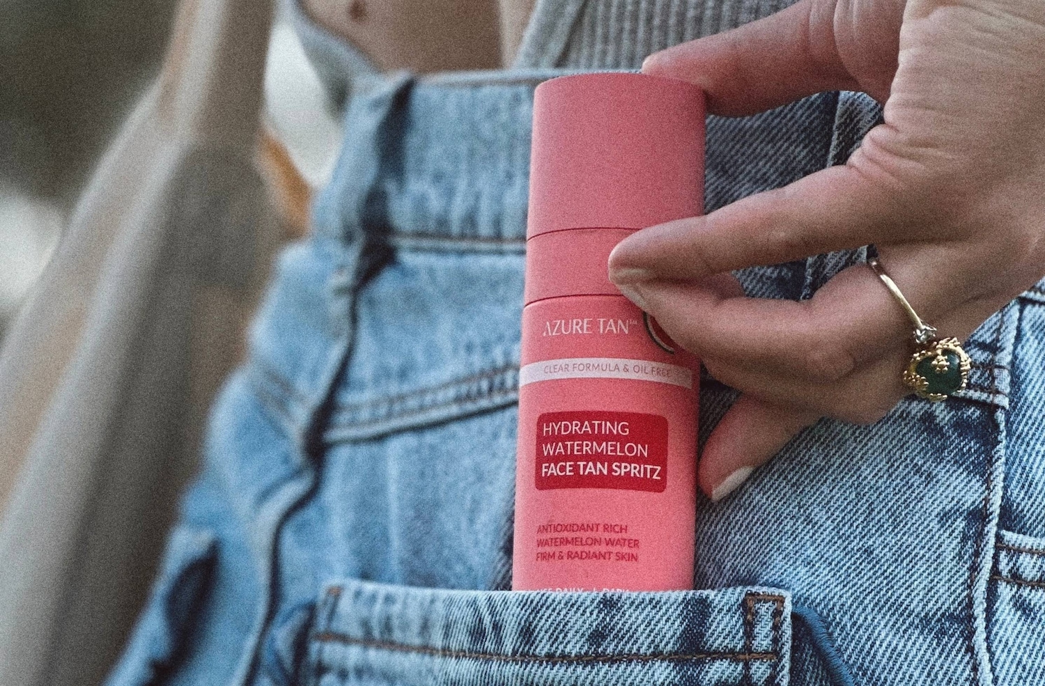 Kobieta trzyma w dłoni różowy spray do twarzy Azure Tan Hydrating Watermelon Face Tan Spritz, który jest umieszczony w kieszeni dżinsów, z widocznym napisem "Clear Formula & Oil Free" oraz "Antioxidant Rich Watermelon Water Firm & Radiant Skin".