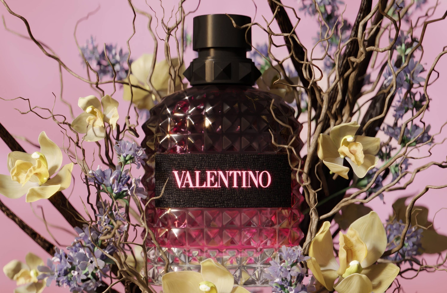 Flakon perfum Valentino w kształcie granatu, z czarną etykietą z napisem „VALENTINO” w kolorze różowym, otoczony jest suchymi gałęziami, jasnożółtymi orchideami i fioletowymi kwiatami, a wszystko to na różowym tle.