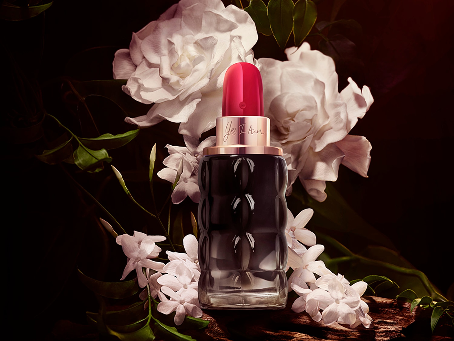 Le parfum "Yes I Am" de Cacharel est présenté dans un flacon en verre noir avec un bouchon rouge vif, entouré de fleurs blanches et de feuilles vertes sur un fond sombre.