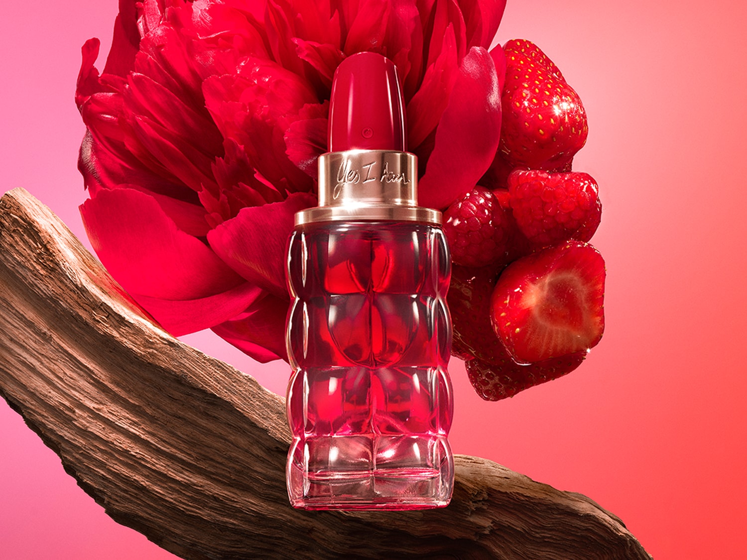 Le parfum Yes I Am de Cacharel, dans un flacon rouge en forme de rouge à lèvres, est posé sur un morceau de bois, entouré de fraises et d'une fleur rouge, sur un fond dégradé de rose et de rouge.