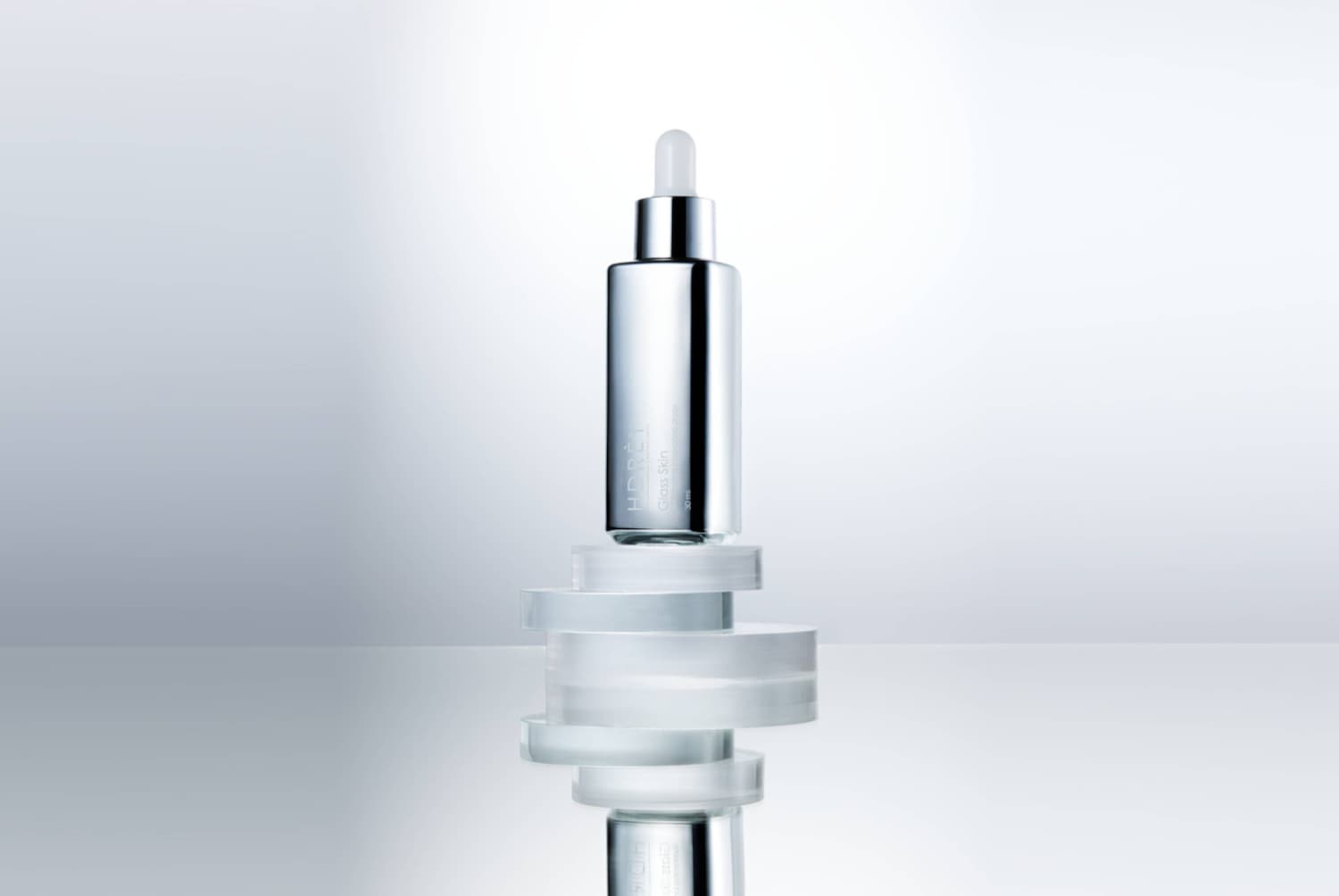 HDRÈY Glass Skin, 30 ml, to serum do twarzy w srebrnej butelce z zakraplaczem, stojące na stosie przezroczystych, okrągłych podkładek, odbijające się w lustrzanej powierzchni.