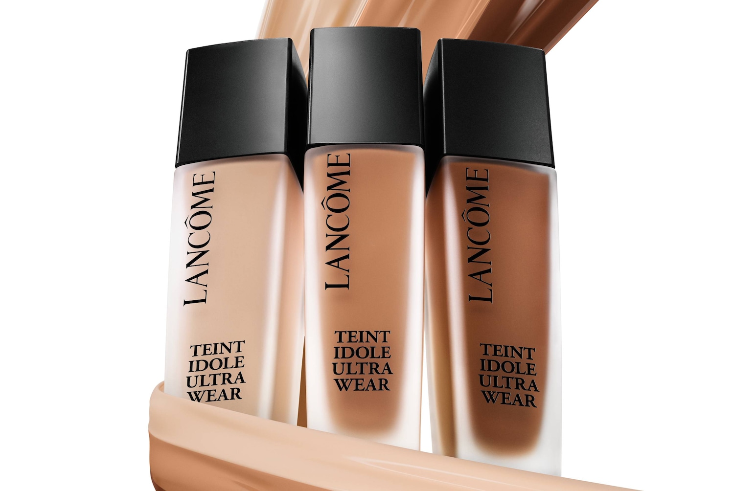 Trzy buteleczki podkładu Lancôme Teint Idole Ultra Wear w odcieniach od jasnego beżu po ciemny brąz, z czarnymi zakrętkami, stoją na tle rozmazanych smug podkładu.