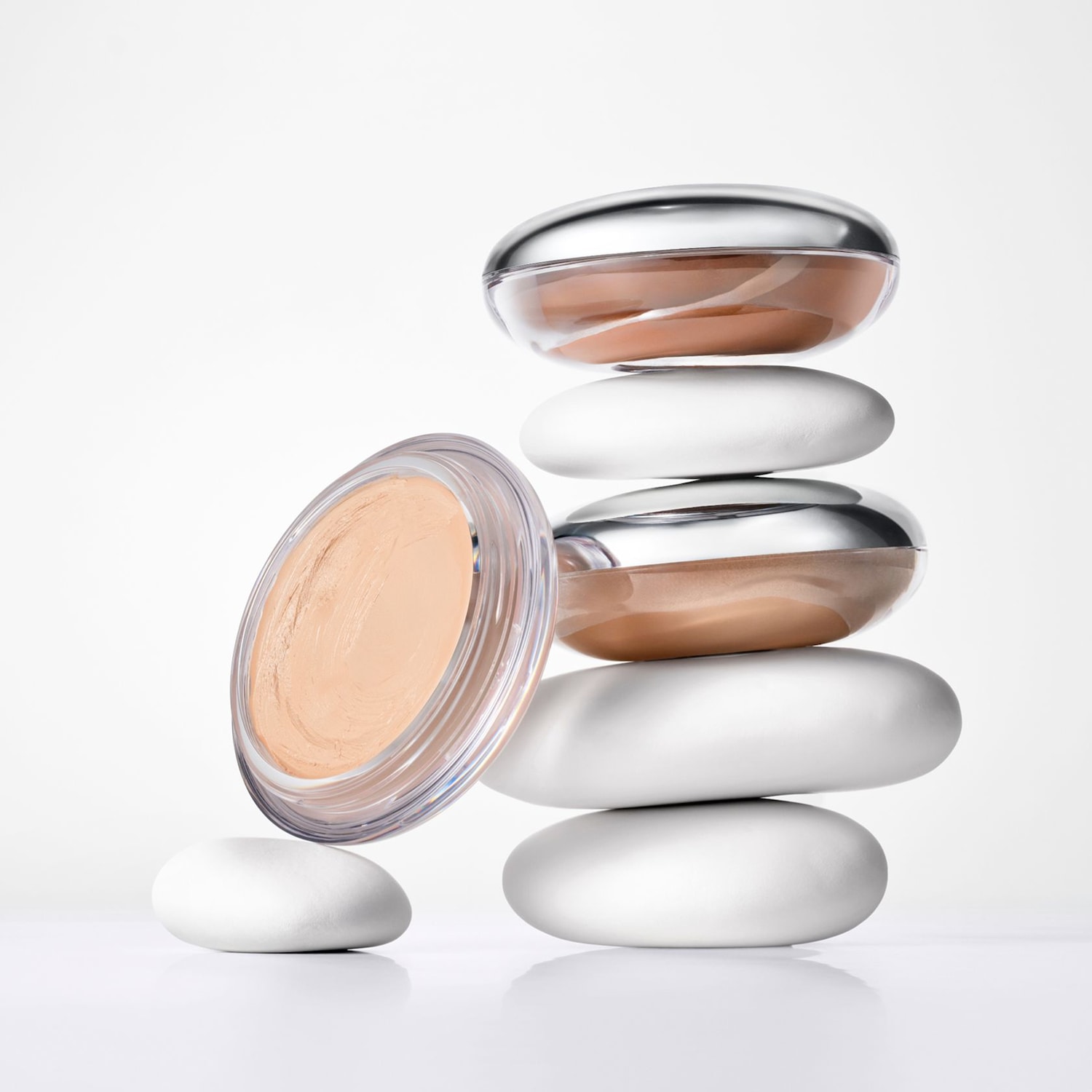 Deux pots de fond de teint compact, l'un de couleur beige clair et l'autre de couleur marron, sont empilés sur des pierres blanches, avec un pot de fond de teint beige clair posé sur le côté.