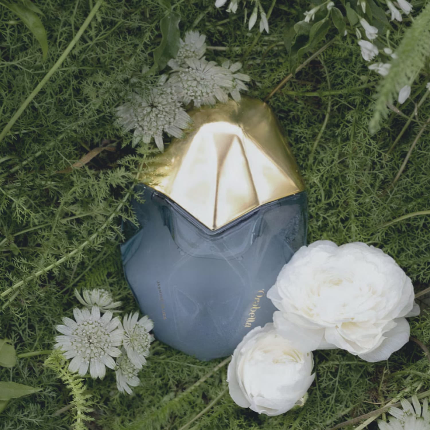 Un flacon de parfum Orabella bleu-gris avec un bouchon doré est posé sur un lit de verdure luxuriante, entouré de délicates fleurs blanches, évoquant une ambiance naturelle et élégante.