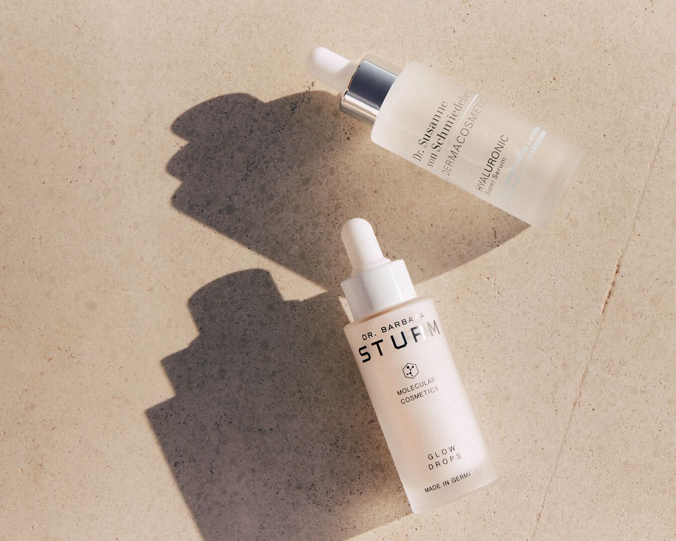 Zwei Hautpflegeseren, Dr. Barbara Sturm Glow Drops und Dr. Susanne von Schmiedeberg Hyaluronic Super Serum, liegen auf einer hellbraunen Oberfläche, bereit, Ihrer Haut einen strahlenden Glanz zu verleihen.