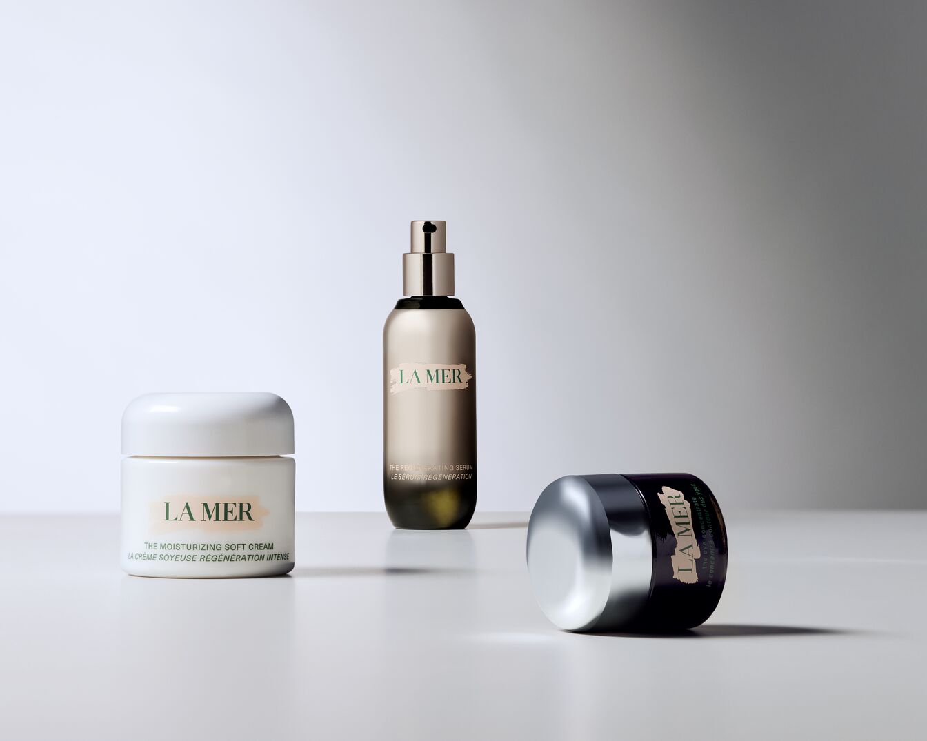 Tři produkty La Mer jsou zobrazeny na bílém pozadí: bílý krém The Moisturizing Soft Cream, zlaté sérum The Regenerating Serum a tmavě fialový oční krém The Eye Concentrate s lesklým stříbrným víčkem.