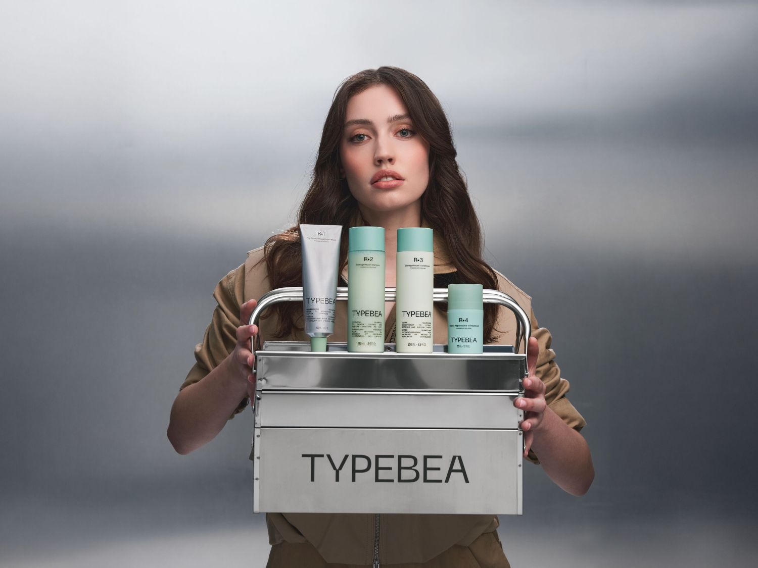 Mladá žena s dlouhými hnědými vlasy drží stříbrný box s nápisem "TYPEBEA", ve kterém jsou čtyři produkty péče o vlasy: stříbrná tuba R+1, světle zelená láhev R+2, světle zelená láhev R+3 a malá světle zelená láhev R+4.