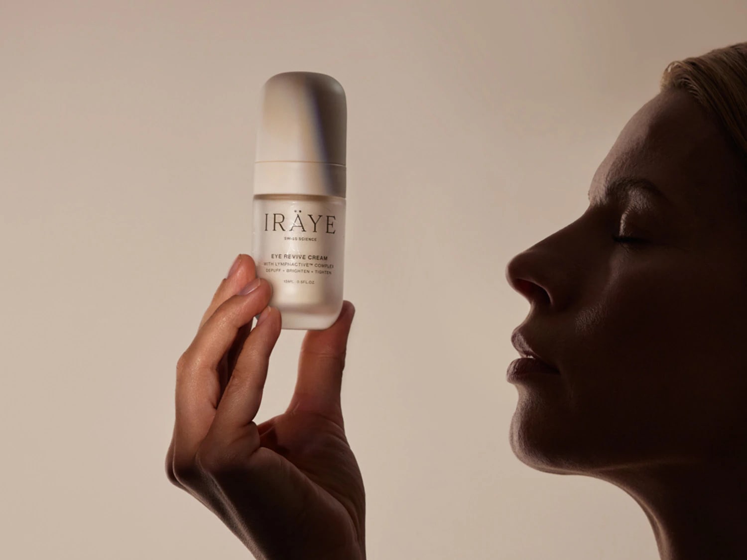 Žena drží v ruce bílý krém na oční okolí IRÄYE Swiss Science Eye Revive Cream s nápisem "depuff, brighten, tighten" a objemem 15 ml, zatímco její obličej je zobrazen z profilu v jemném světle.