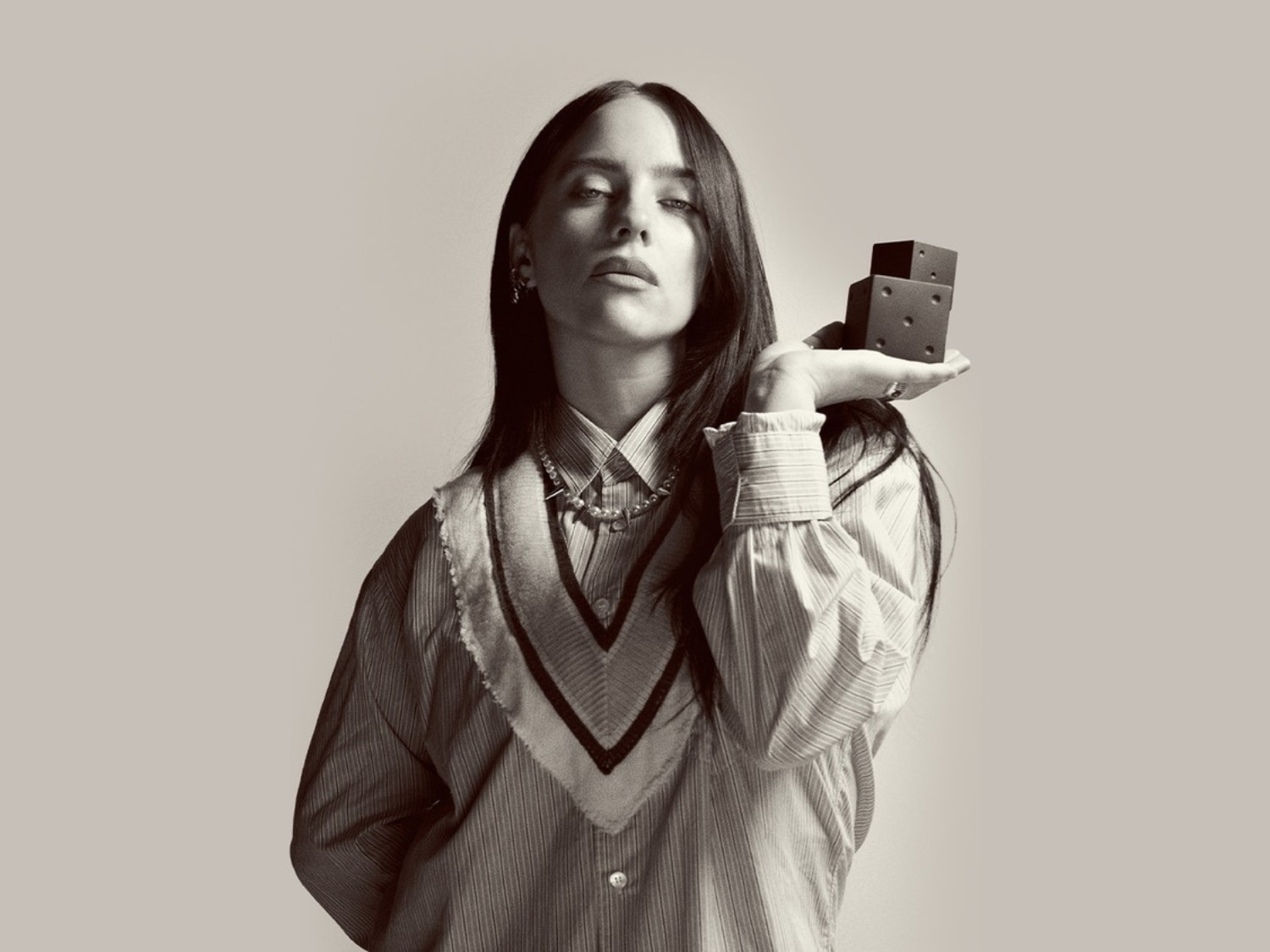 Billie Eilish pózuje s černými kostkami v ruce, má na sobě pruhovanou košili a svetr s výstřihem do V, což podtrhuje její jedinečný styl.