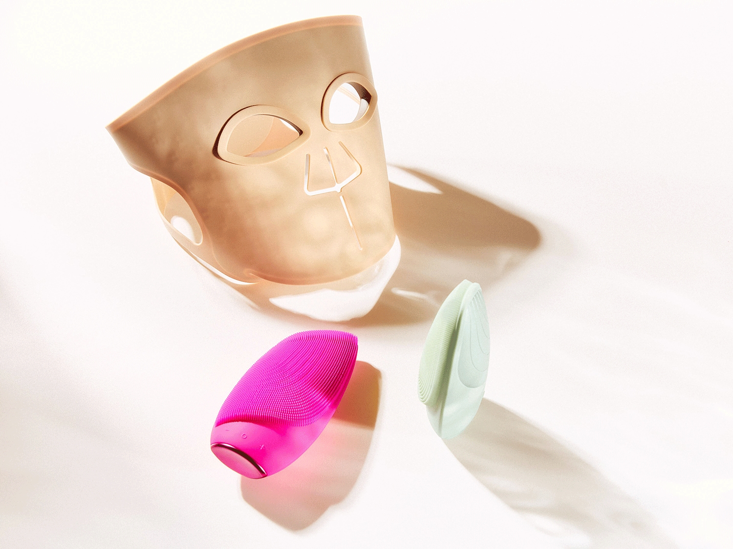 Ein hellbraunes Gesichtsmasken-Gerät mit Aussparungen für Augen, Nase und Mund liegt neben zwei ovalen Reinigungsbürsten in leuchtendem Pink und Hellgrün auf einem weißen Hintergrund.