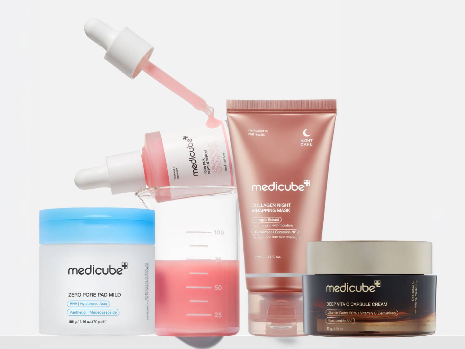 Zestaw produktów Medicube, w tym serum peptydowe PDRN w różowej butelce z zakraplaczem, maska na noc Collagen Night Wrapping Mask w tubce w kolorze różowego złota, krem Deep Vita C Capsule Cream w ciemnym słoiczku oraz płatki Zero Pore Pad Mild w białym pojemniku z niebieską pokrywką, wszystkie ułożone na tle szklanej zlewki z różowym płynem.