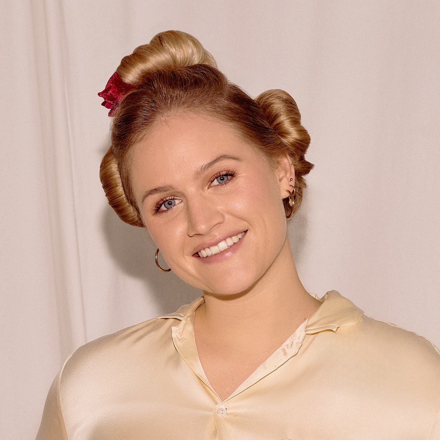 Eine lächelnde Frau mit blonden Haaren, die zu zwei Dutts hochgesteckt sind, trägt ein rotes Haarband und goldene Creolen, während sie ein cremefarbenes Hemd anhat.